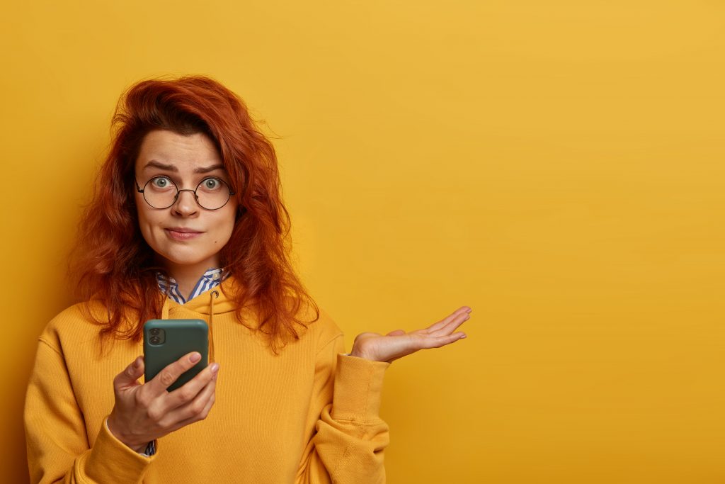 indignant-puzzled-redhead-woman-raises-palm-thinks-what-to-answer-on-received-message-holds-mobile-phone-wears-round-spectacles-and-hoodie-models-over-yellow-wall-with-blank-space-right-scaled - HERRAMIENTAS DE CONTROL PARENTAL: SEGURIDAD EN LA RED
